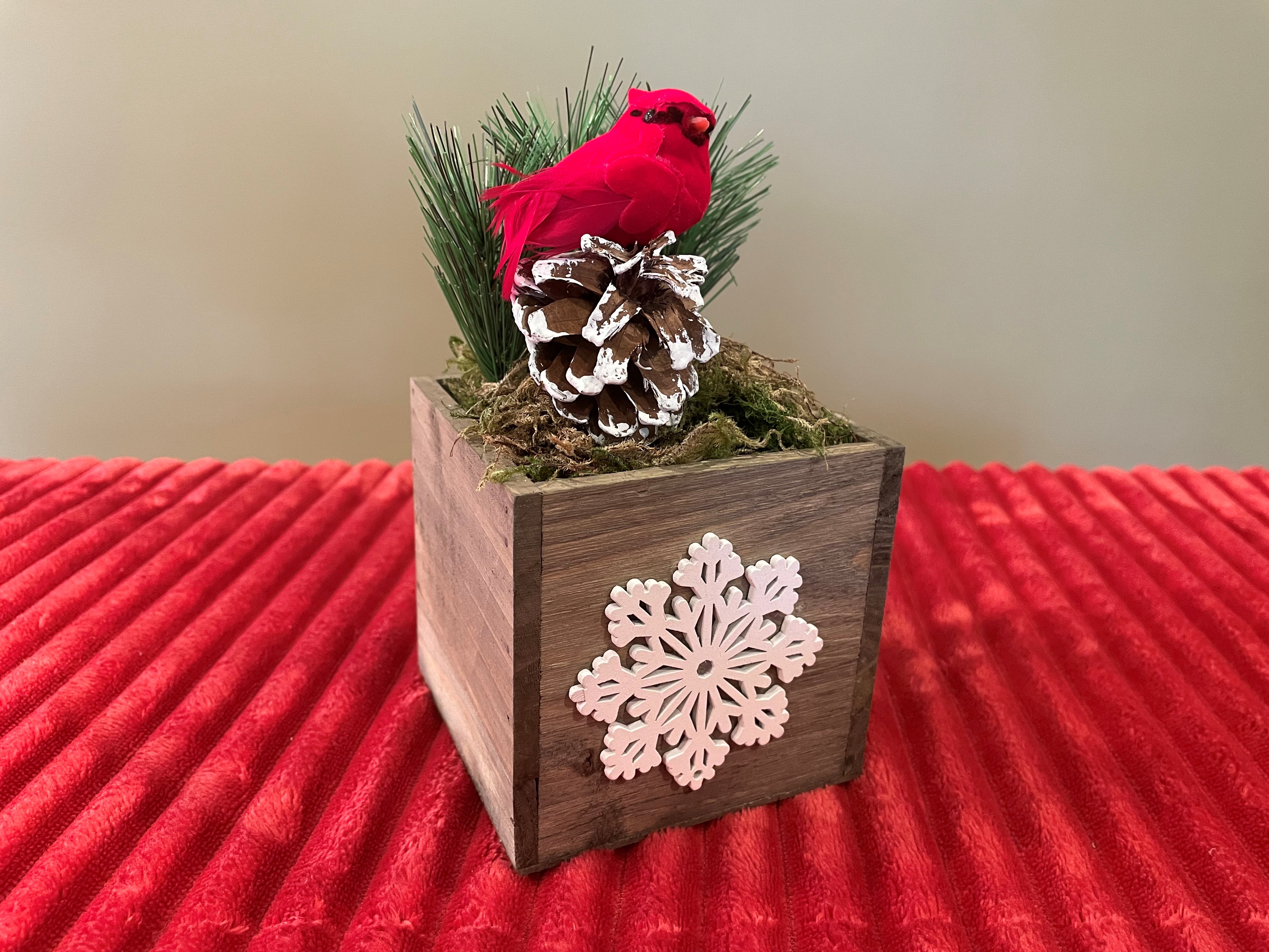 A centerpiece made of a wooden planter with a white snowflake on the front. Filled with greenery, a pine cone, and a red cardinal bird.