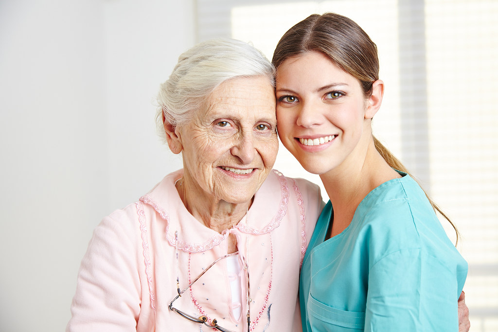 An elderly white woman in a pink dressing gown stands closely to a younger white woman wearing aqua-colored medical scrubs.