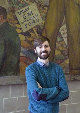 A photograph of UAW Archivist Gavin Strassel, standing in front of a labor mural with his arms crossed.