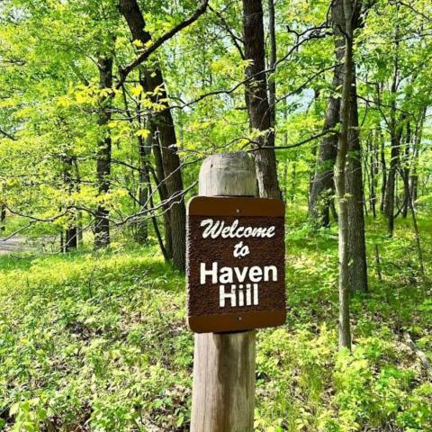 A color photo of a brown historic place sign marker reading, "Welcome to Haven Hill". It is standing in a forest of trees.