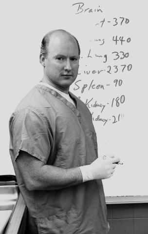 A black and white photography of author Tobin Buhk in scrubs and gloves, standing in front of a white board.