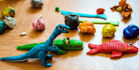 A blue dinosaur, red fish, and green alligator made of Play-Doh are surrounded by balls of Play-Doh and tools for sculpting.